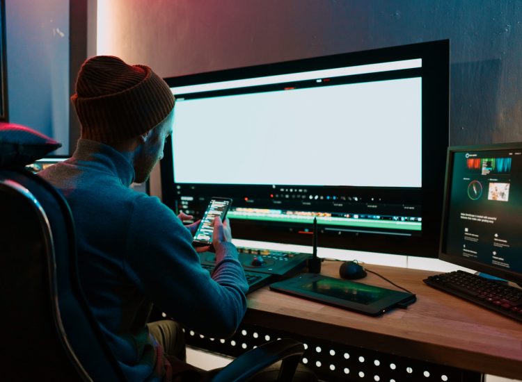 Attractive Male Video Editor Works with Footage or Video on His Personal Computer and having a break communicating on his smartphone. He Works in Creative Office Studio or home. Neon lights